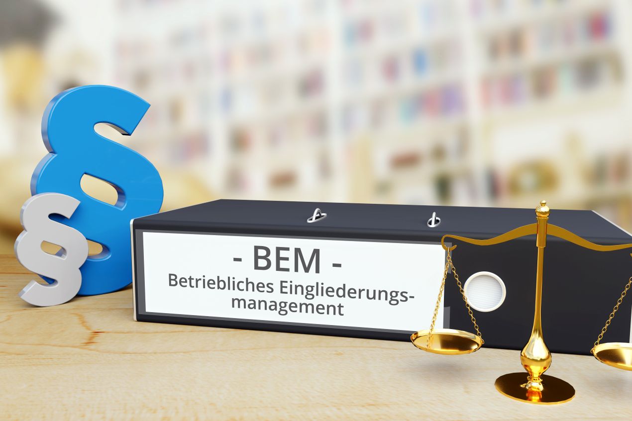 Schwarzer Aktenordner mit Aufschrift "BEM – Betriebliches Eingliederungsmanagement", daneben ein Paragraphensymbol und eine goldene Waage als Symbol für rechtliche Aspekte.