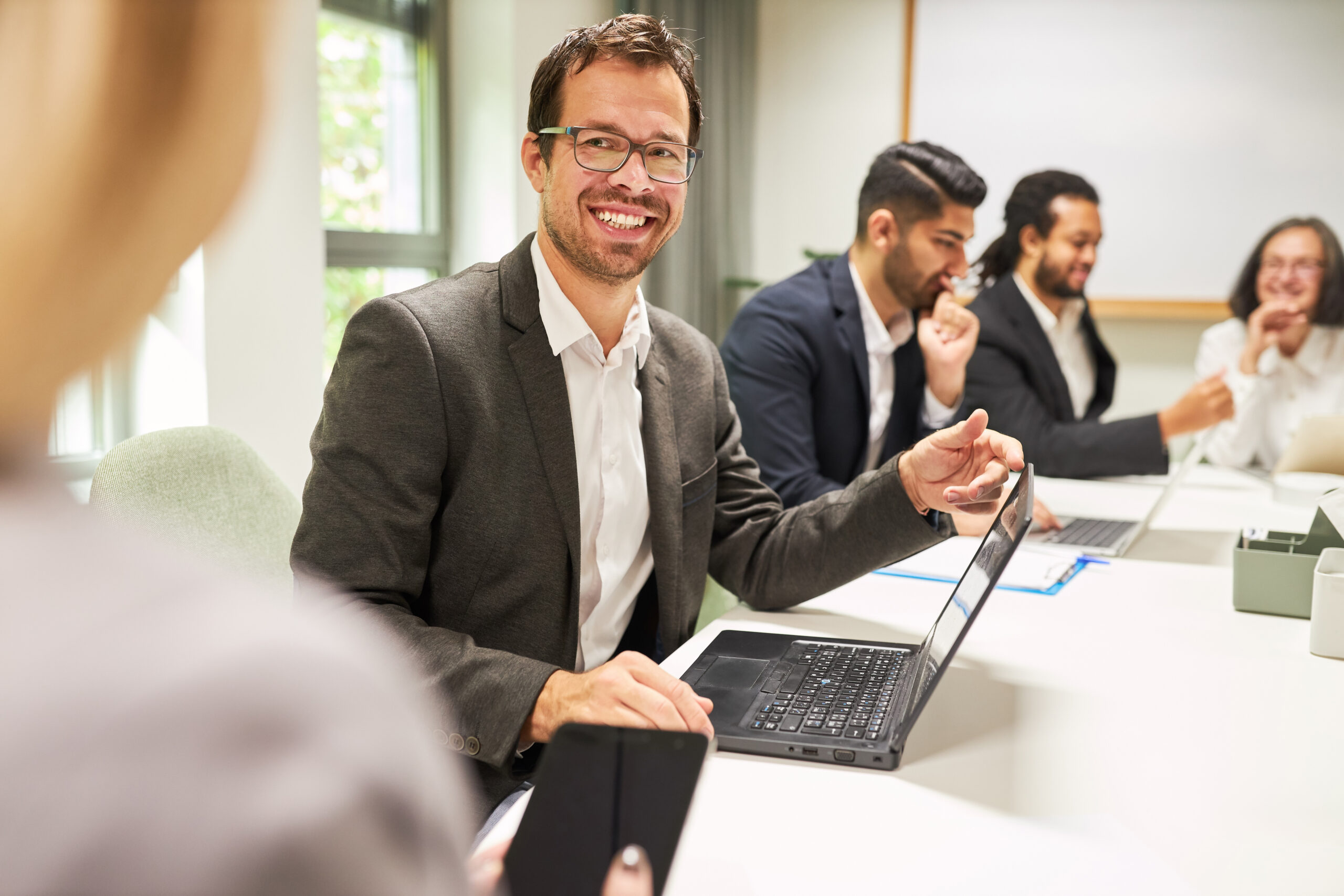 Lächelnder Mann in Business-Outfit sitzt mit Laptop in einer Teambesprechung, umgeben von Kollegen – steht für die konstruktive Umsetzung des BEM-Verfahrens.