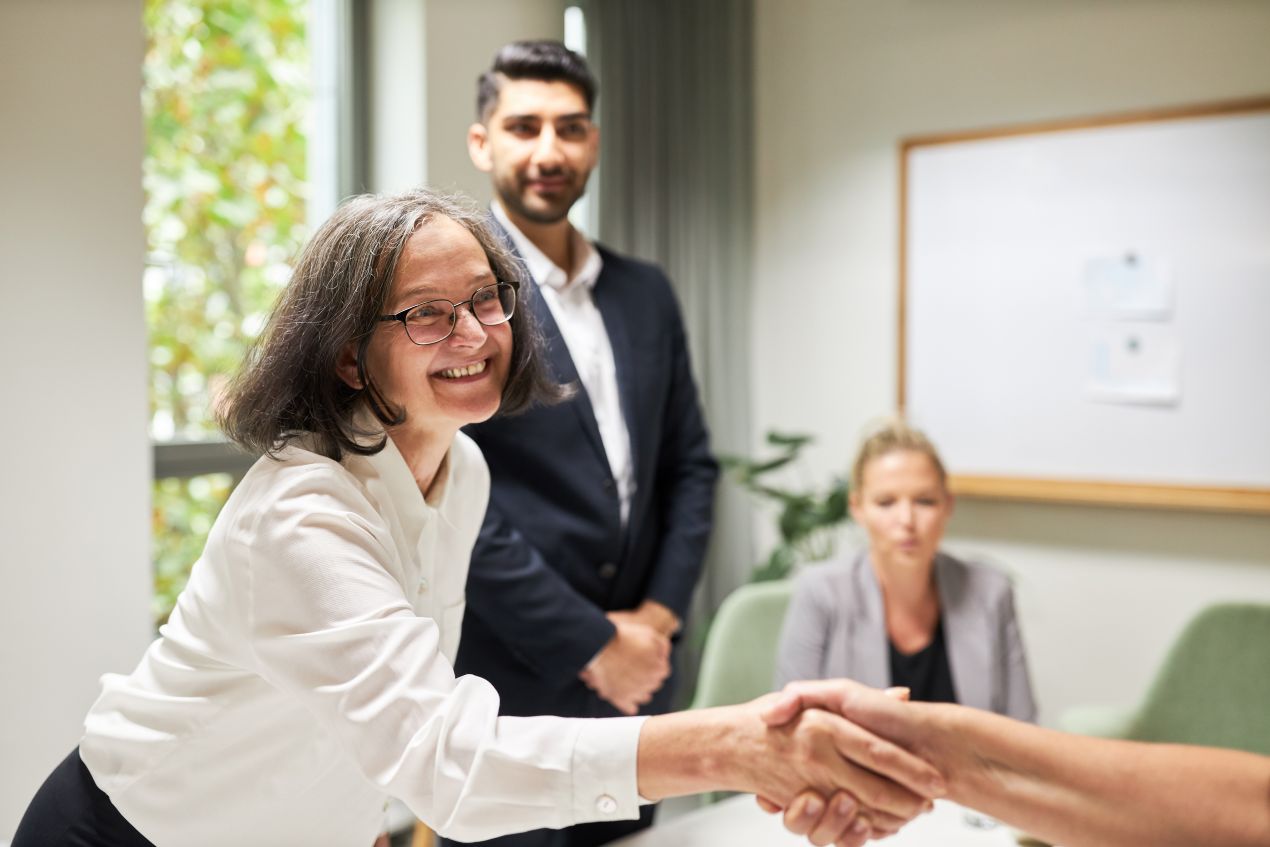 Freundliche Frau in Business-Kleidung schüttelt die Hand eines Gesprächspartners in einem Büro, während zwei Kollegen im Hintergrund beobachten – Symbol für ein gelungenes BEM-Gespräch.